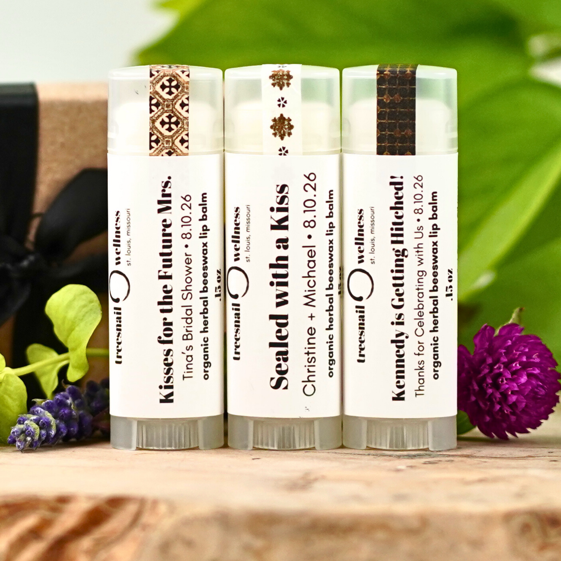 Three lip balm sticks with personalized text on a wooden surface with flowers in the background