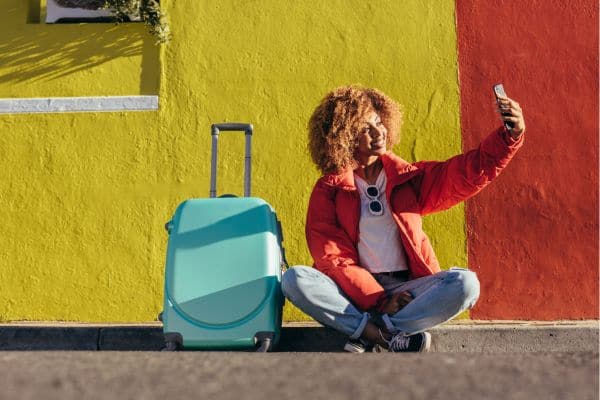a woman taking a selfie in front of a bright yellow and red concrete wall next to a turquoise suitcase