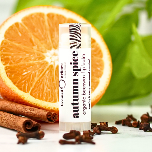 Lip balm labeled 'autumn spice' with an orange, cinnamon sticks, and cloves in the background.