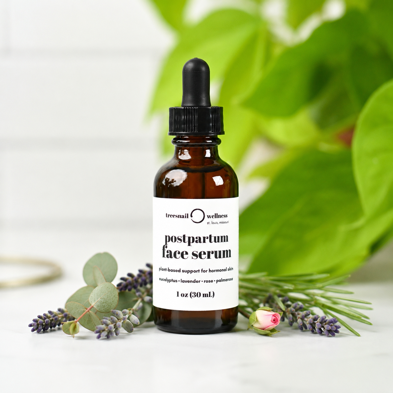An amber glass dropper bottle of Tressail Wellness postpartum face serum, styled with eucalyptus leaves, lavender sprigs, and a rose bud on a white surface with a soft-focus green plant background.