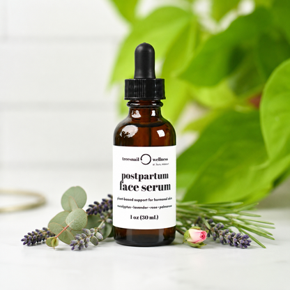 An amber glass dropper bottle of Tressail Wellness postpartum face serum, styled with eucalyptus leaves, lavender sprigs, and a rose bud on a white surface with a soft-focus green plant background.