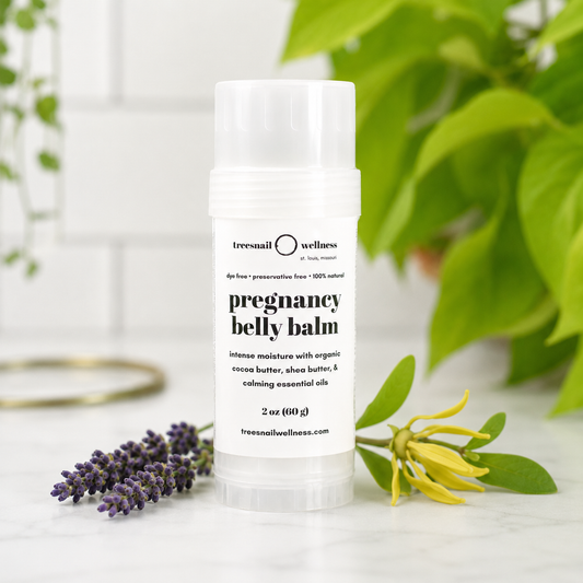 A 2 oz stick of Tressaith Wellness Pregnancy Belly Balm standing upright on a marble surface, surrounded by green leaves, a purple lavender sprig, and a yellow flower.