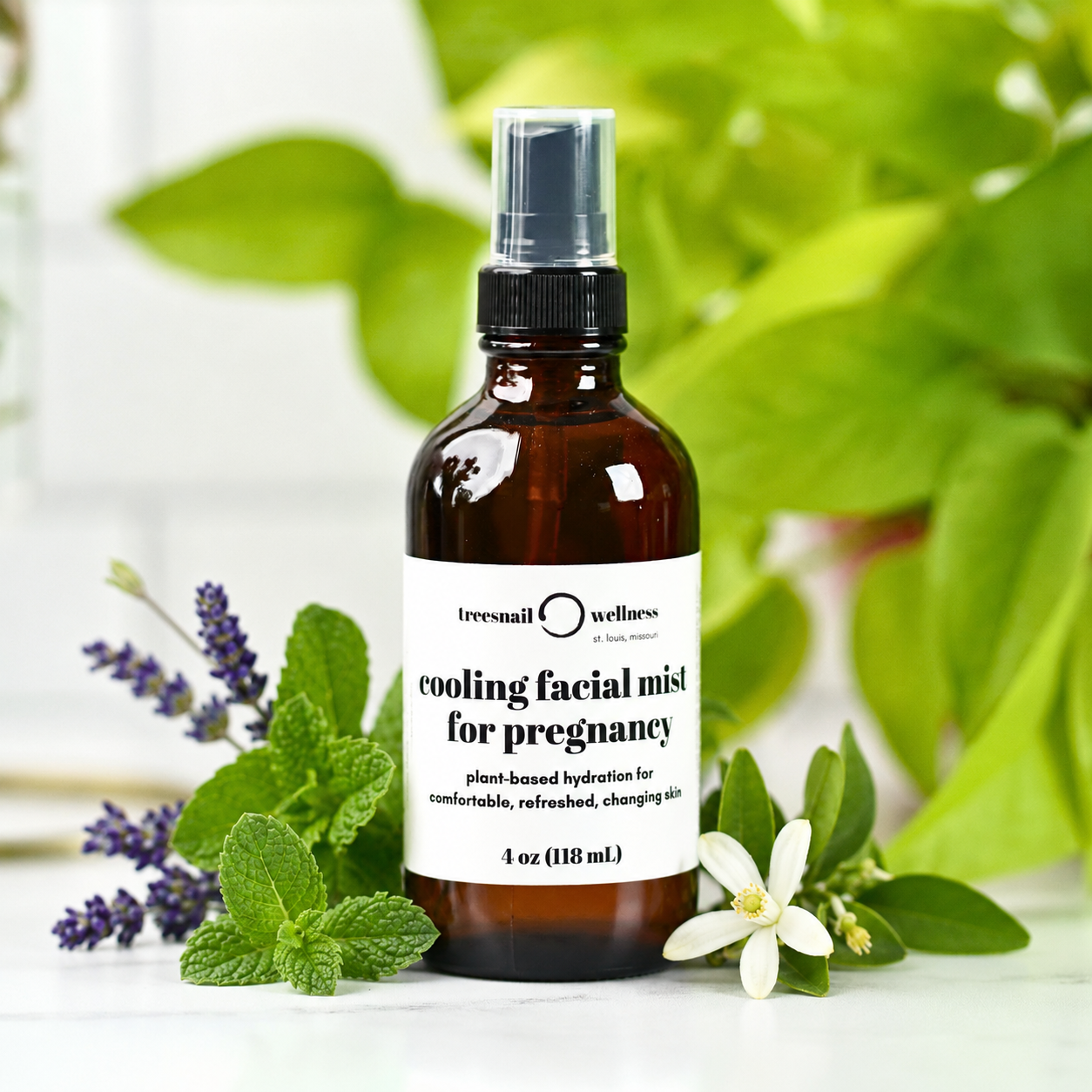 A 4 oz amber glass spray bottle of Tressnail Wellness Cooling Facial Mist for Pregnancy, styled with fresh green leaves, mint, and a white flower on a light surface.