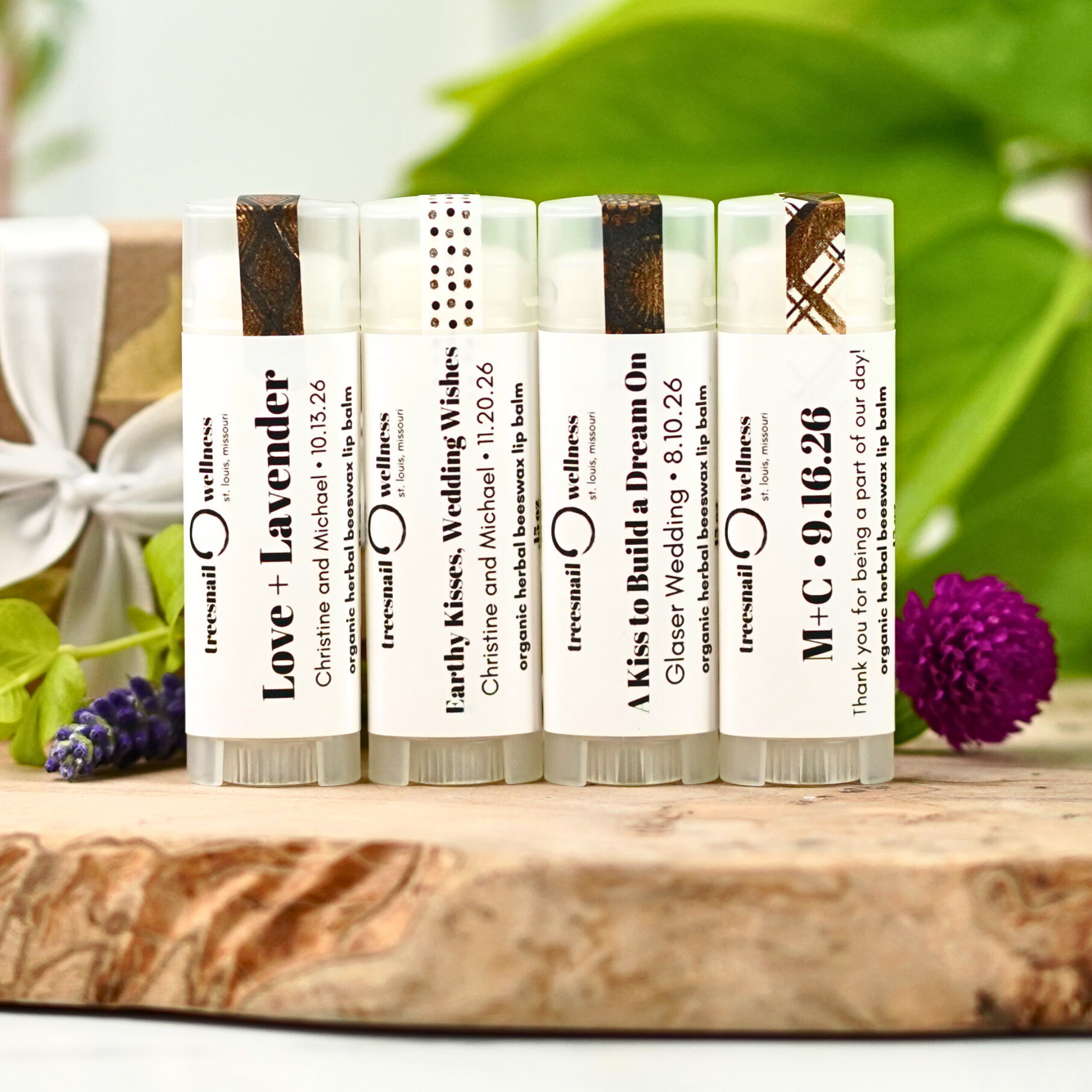 Set of wedding favor lip balms with personalized text on a wooden surface with greenery in the background