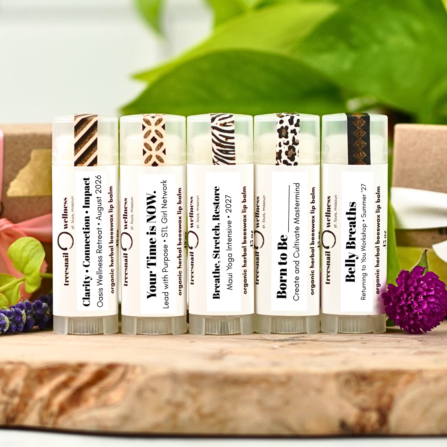 Set of custom lip balms with labels on a wooden surface with greenery in the background